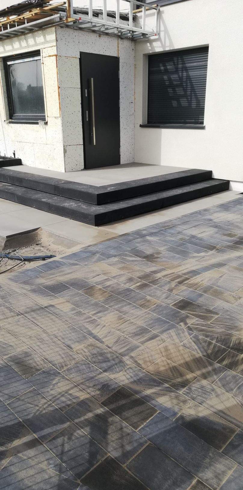 Nowo ułożona, wielobarwna kostka brukowa prowadząca do wejścia z dwoma stopniami wykończonymi czarnym obrzeżem, widoczny fragment elewacji z oknami.