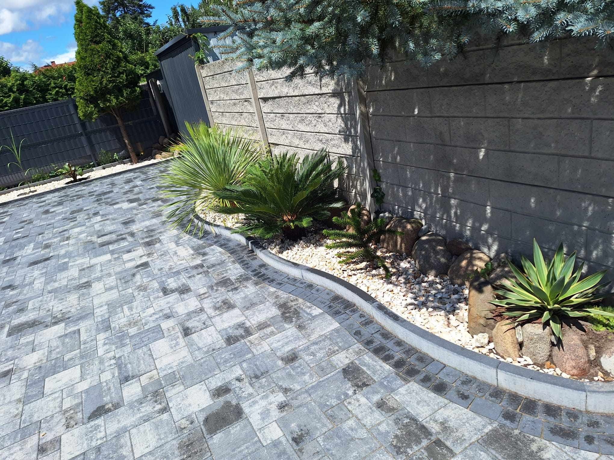 Ułożona szara kostka brukowa w geometryczny wzór, obrzeże z kostki, rabata z kamieniami, egzotycznymi roślinami i korą pod betonowym murem w ogrodzie.