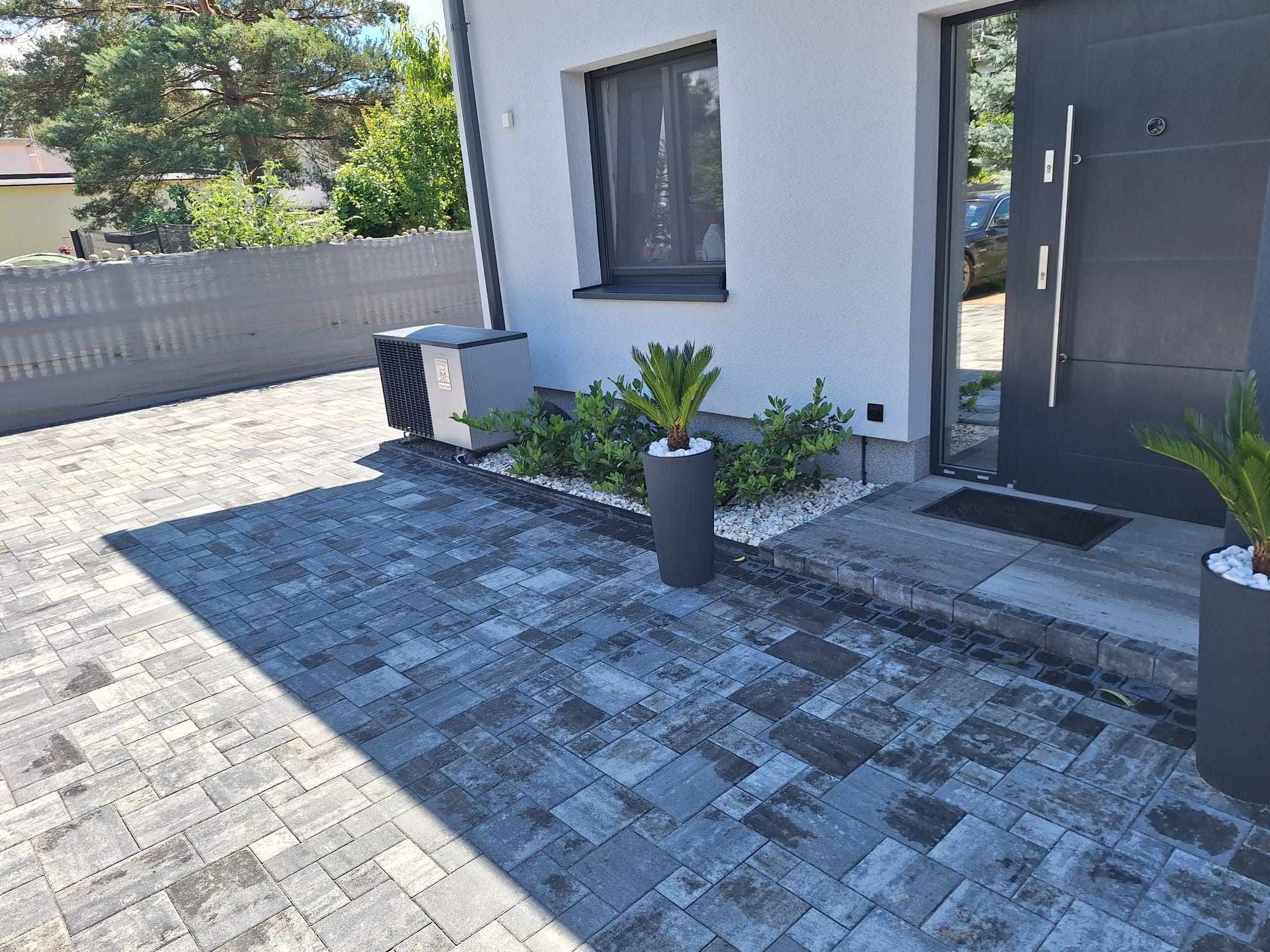 Ułożona szara kostka brukowa w różnych odcieniach szarości prowadząca do nowoczesnego domu z szarymi drzwiami i oknem, z roślinami w donicach i klimatyzatorem na zewnątrz