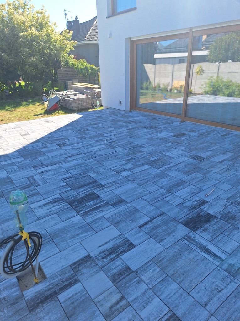 Nowo ułożony taras z szarej kostki o nieregularnym wzorze, z widocznym w tle domem z dużym oknem tarasowym i stertą kostki, wężyk z wodą do zwilżania kostki leży na ziemi.