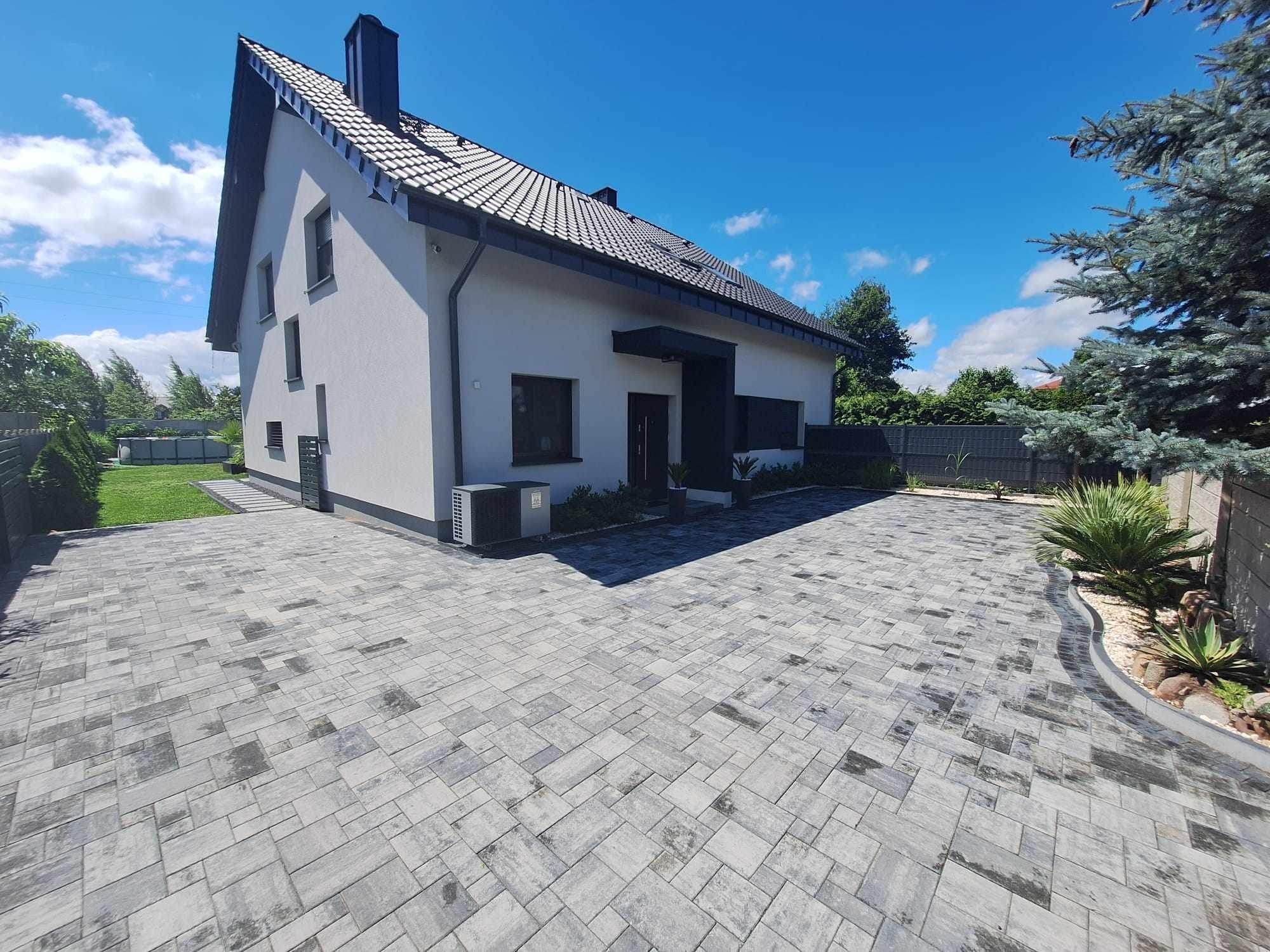 Nowoczesny dom jednorodzinny z szarą kostką brukową ułożoną we wzór prostokątów, widok z niskiej perspektywy podkreślający dużą powierzchnię podjazdu i harmonijne połączenie z architekturą budynku.