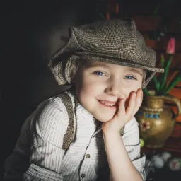 Portret uśmiechniętego chłopca w kaszkiecie i szelkach, opierającego policzek na dłoni, z dekoracyjną wazą z tulipanami i koszykiem w tle.