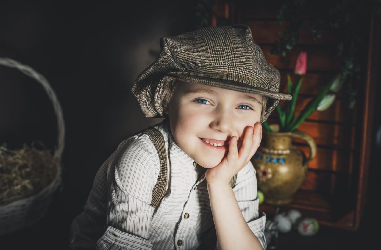 Portret uśmiechniętego chłopca w kaszkiecie i szelkach, opierającego policzek na dłoni, z dekoracyjną wazą z tulipanami i koszykiem w tle.