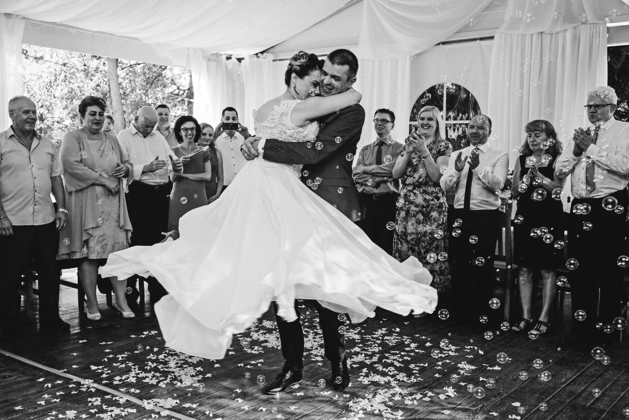 Czarno-białe zdjęcie pary młodej tańczącej w otoczeniu gości podczas wesela, z bańkami mydlanymi w powietrzu i konfetti na podłodze.
