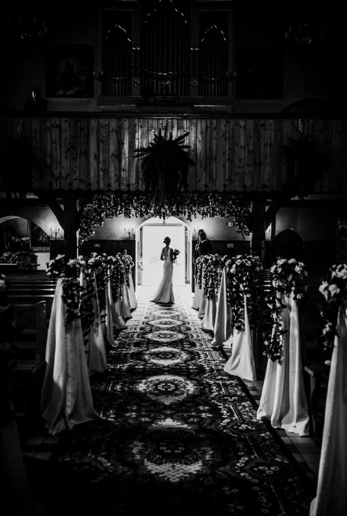 Czarno-białe zdjęcie panny młodej w sukni ślubnej wchodzącej do kościoła, z dekoracjami kwiatowymi i materiałowymi przy ławkach oraz wzorzystym dywanem na posadzce, widok od tyłu, zarys organów...