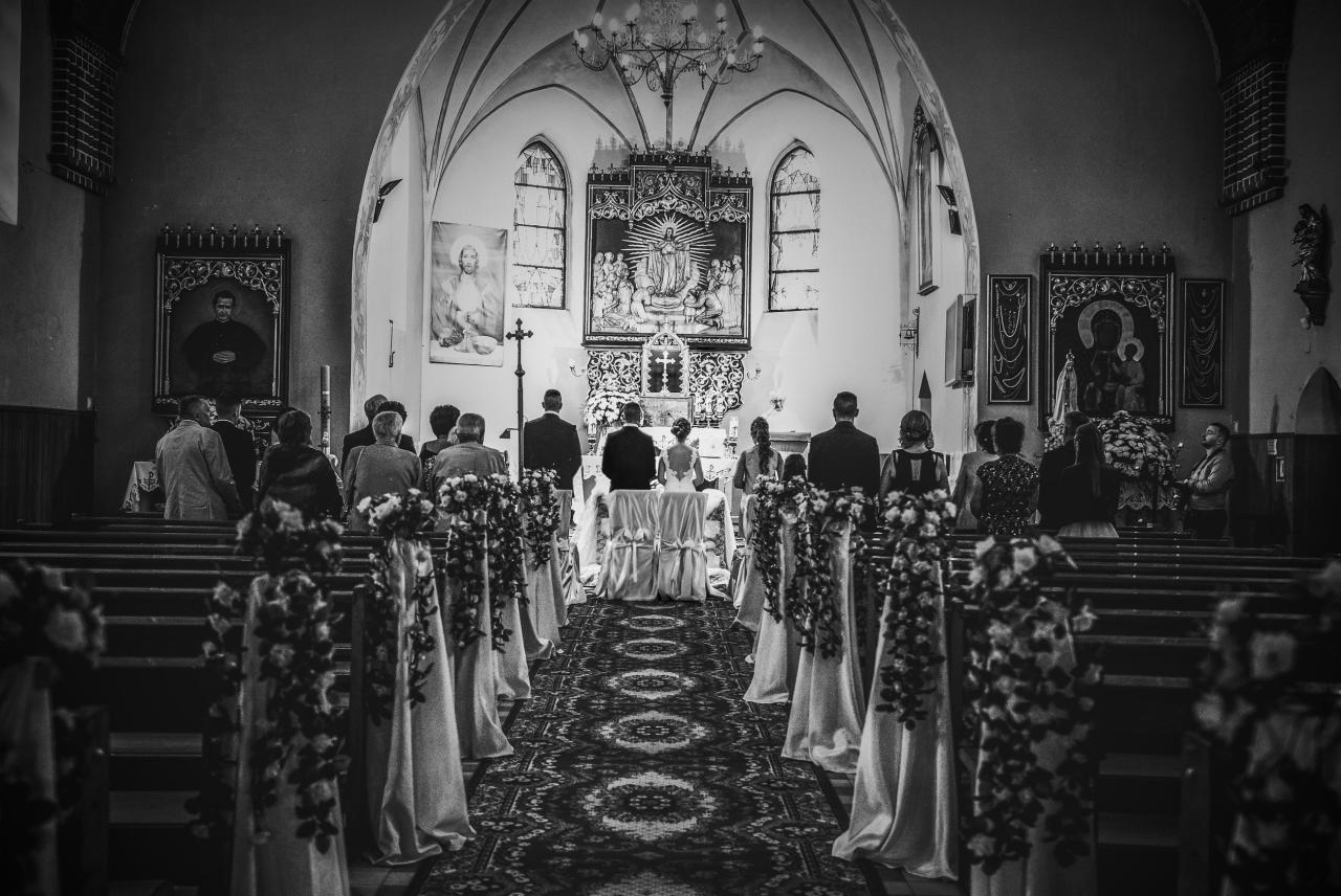 Czarno-białe zdjęcie wnętrza kościoła podczas ceremonii ślubnej, z parą młodą stojącą przed ołtarzem, widoczne dekoracje kwiatowe na ławkach i goście siedzący w ławkach.