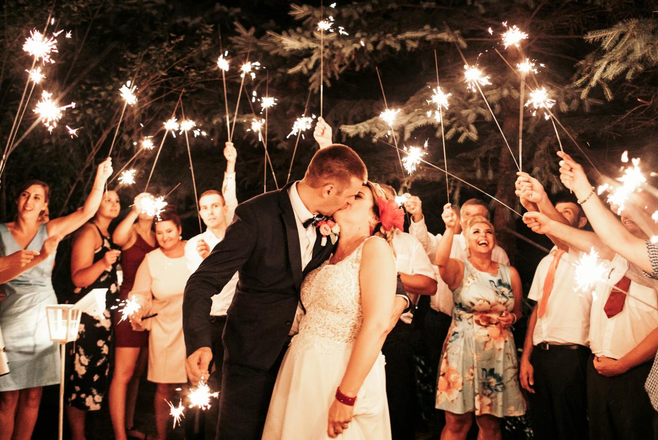 Pocałunek pary młodej otoczonej gośćmi weselnymi z zimnymi ogniami w wieczornej scenerii.