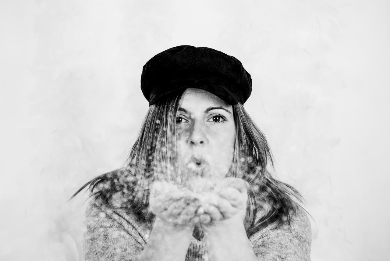 Czarno-białe zbliżenie kobiety w berecie dmuchającej brokat z dłoni, z widocznymi drobinkami w powietrzu na rozmytym tle.