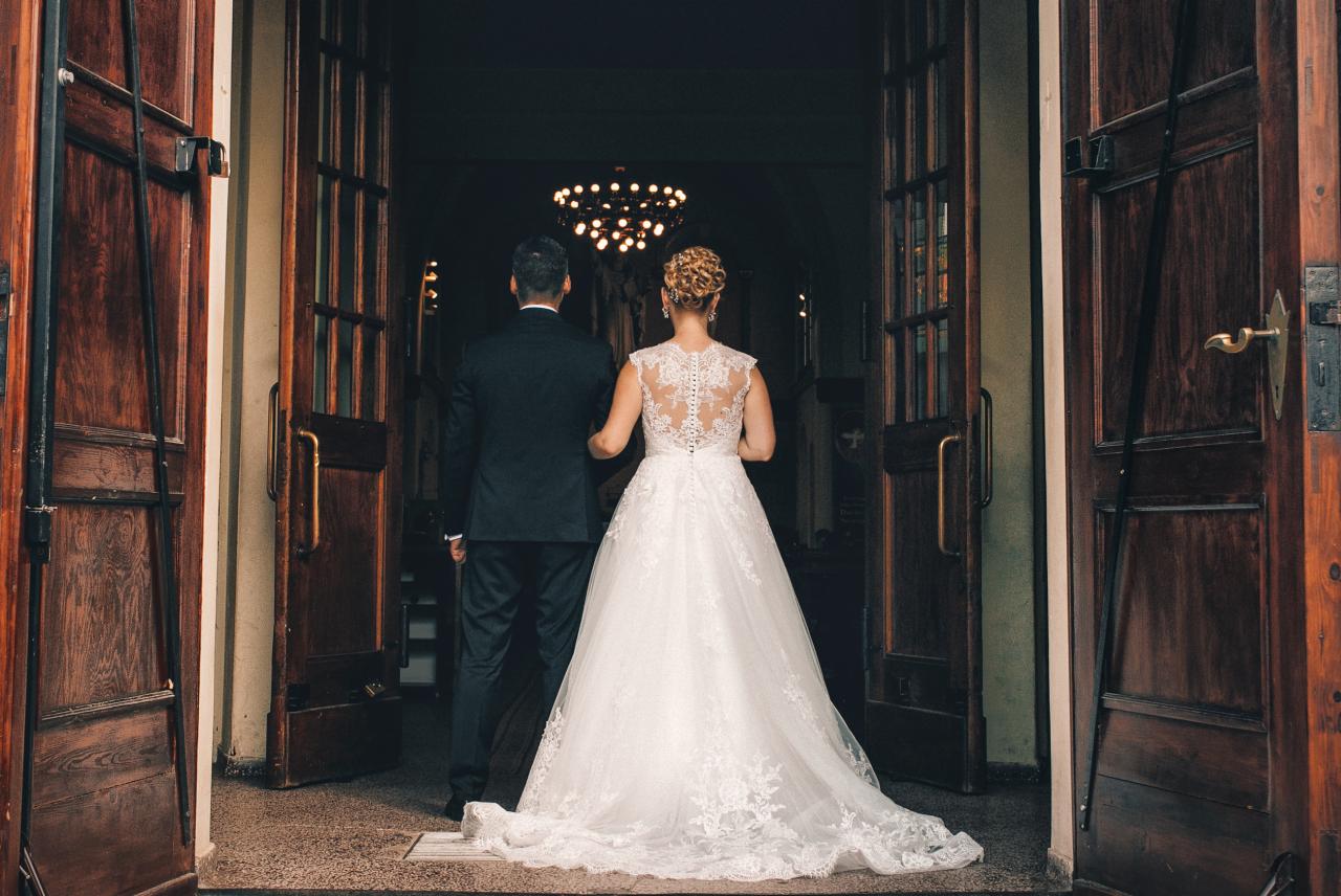 Para młoda wchodzi do kościoła przez otwarte, drewniane drzwi. Suknia panny młodej z koronkowymi detalami na plecach. W tle widoczny żyrandol.