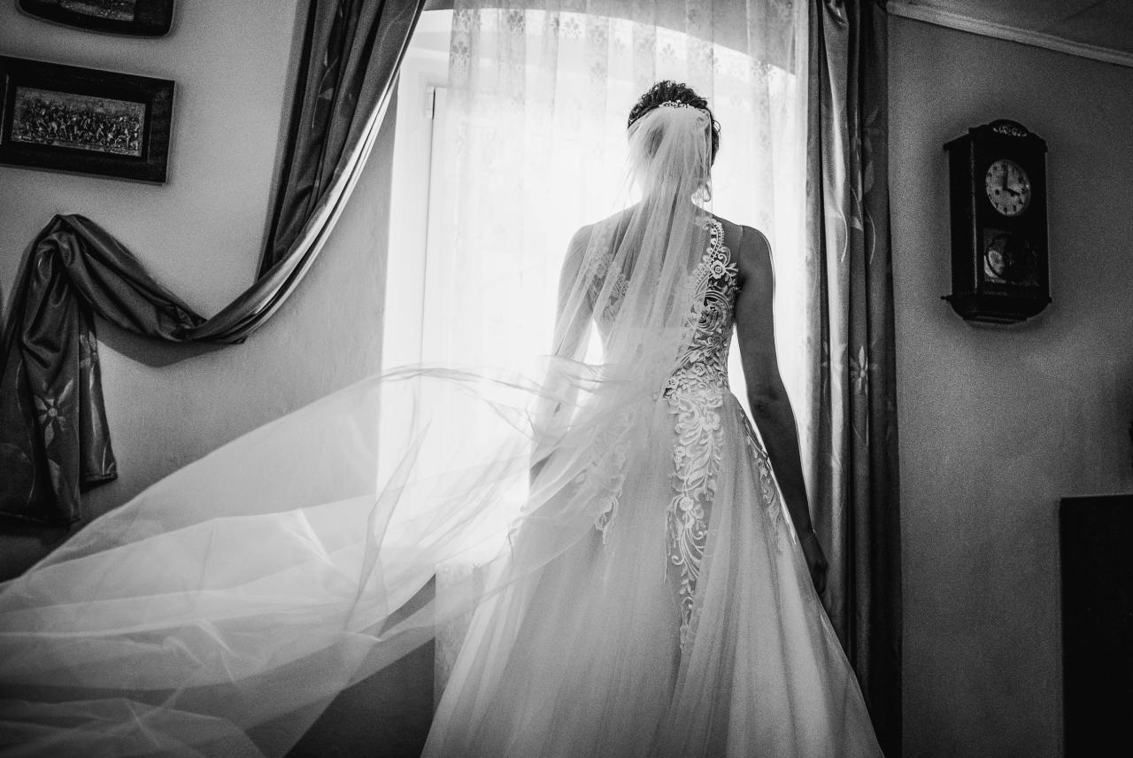 Panna młoda w koronkowej sukni i welonie, stojąca tyłem do okna, z powiewającym welonem, w tle zegar ścienny i dekoracja ścienna.