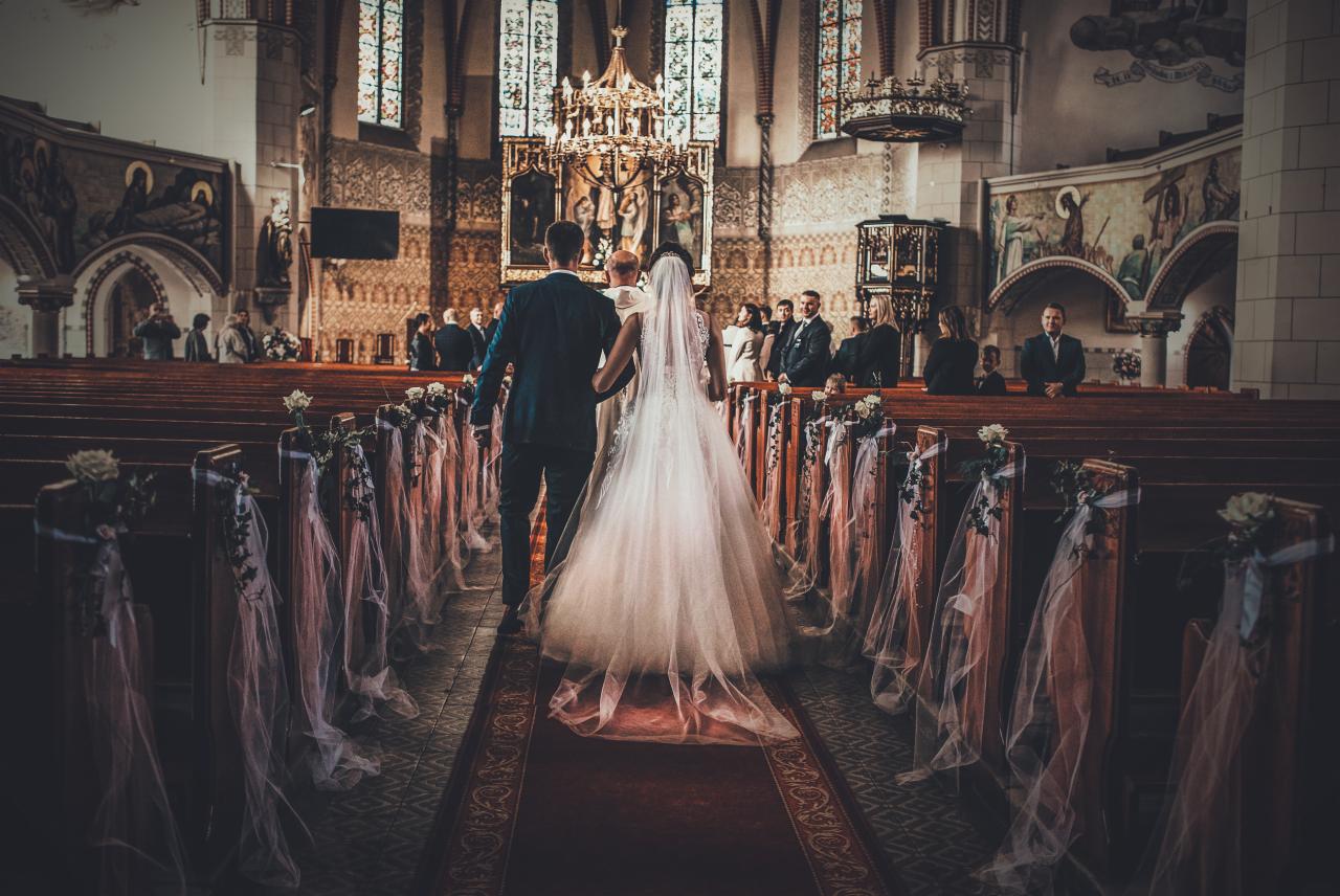 Para młoda idzie przez kościół udekorowany białymi kwiatami i tiulem wzdłuż ławek, w tle goście i ołtarz.