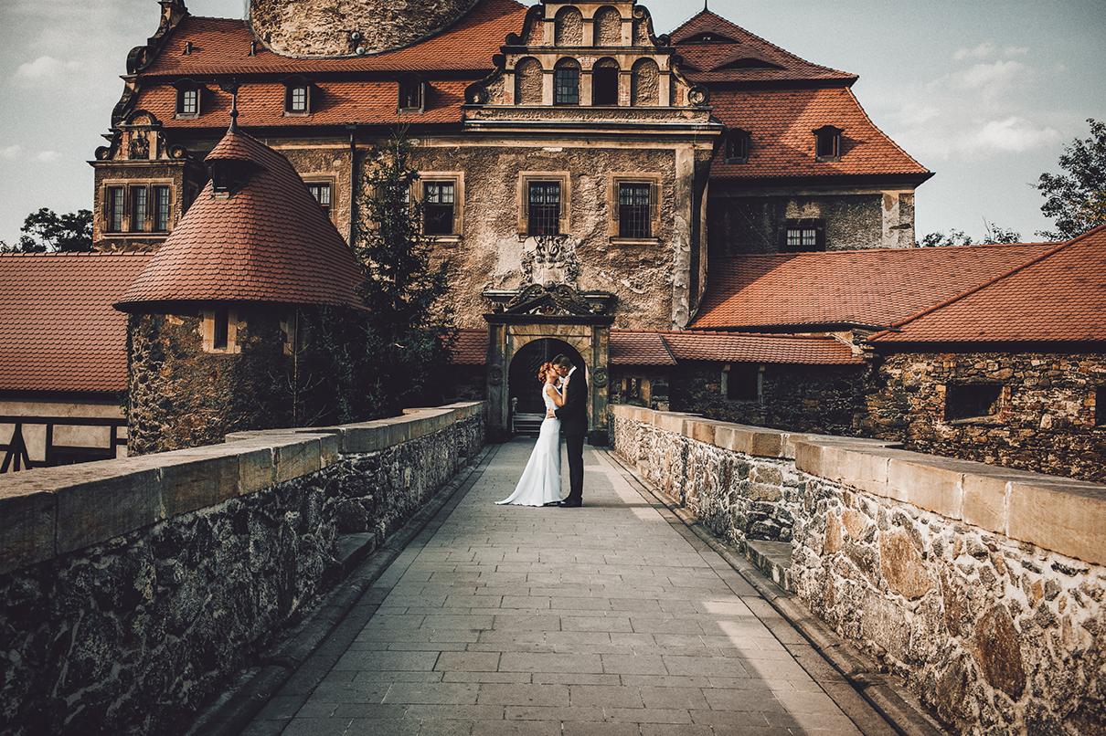 Para młoda w objęciach na kamiennym moście prowadzącym do zamku z czerwonymi dachami i ozdobnym portalem wejściowym.