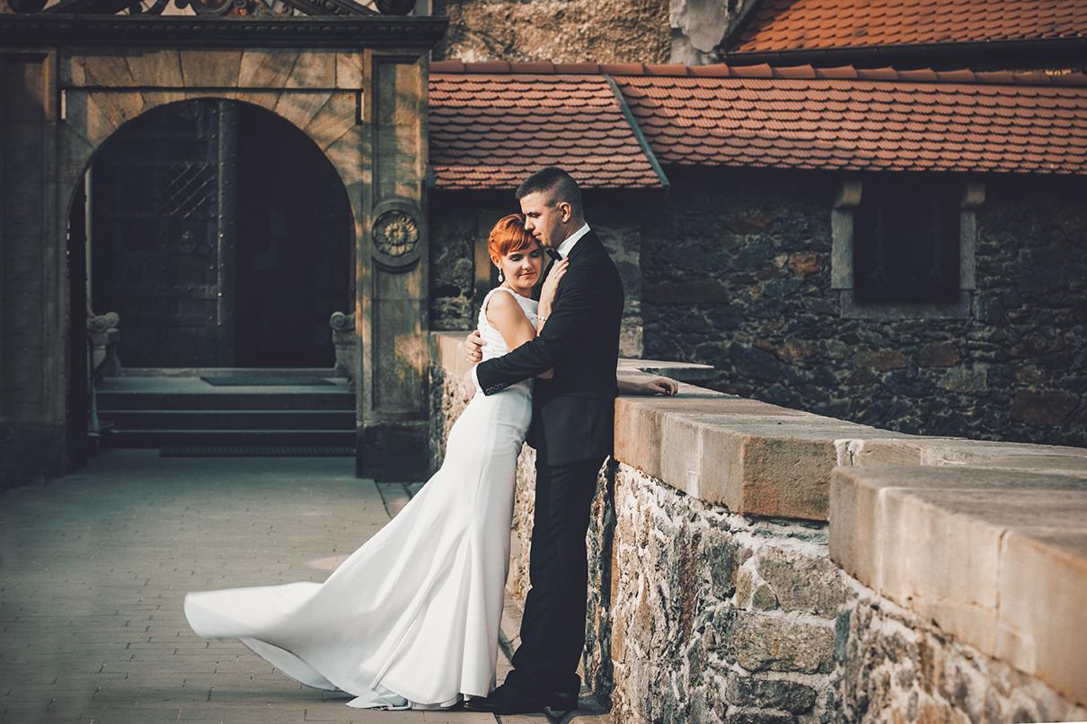 Para młoda przytulona na tle kamiennego zamku z czerwoną dachówką, panna młoda w białej sukni z trenem, pan młody w czarnym garniturze.