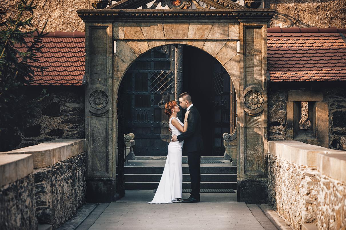 Para młoda w uścisku na tle kamiennej bramy zamku, panna młoda w białej sukni z odkrytymi ramionami, pan młody w czarnym garniturze, romantyczna sceneria.