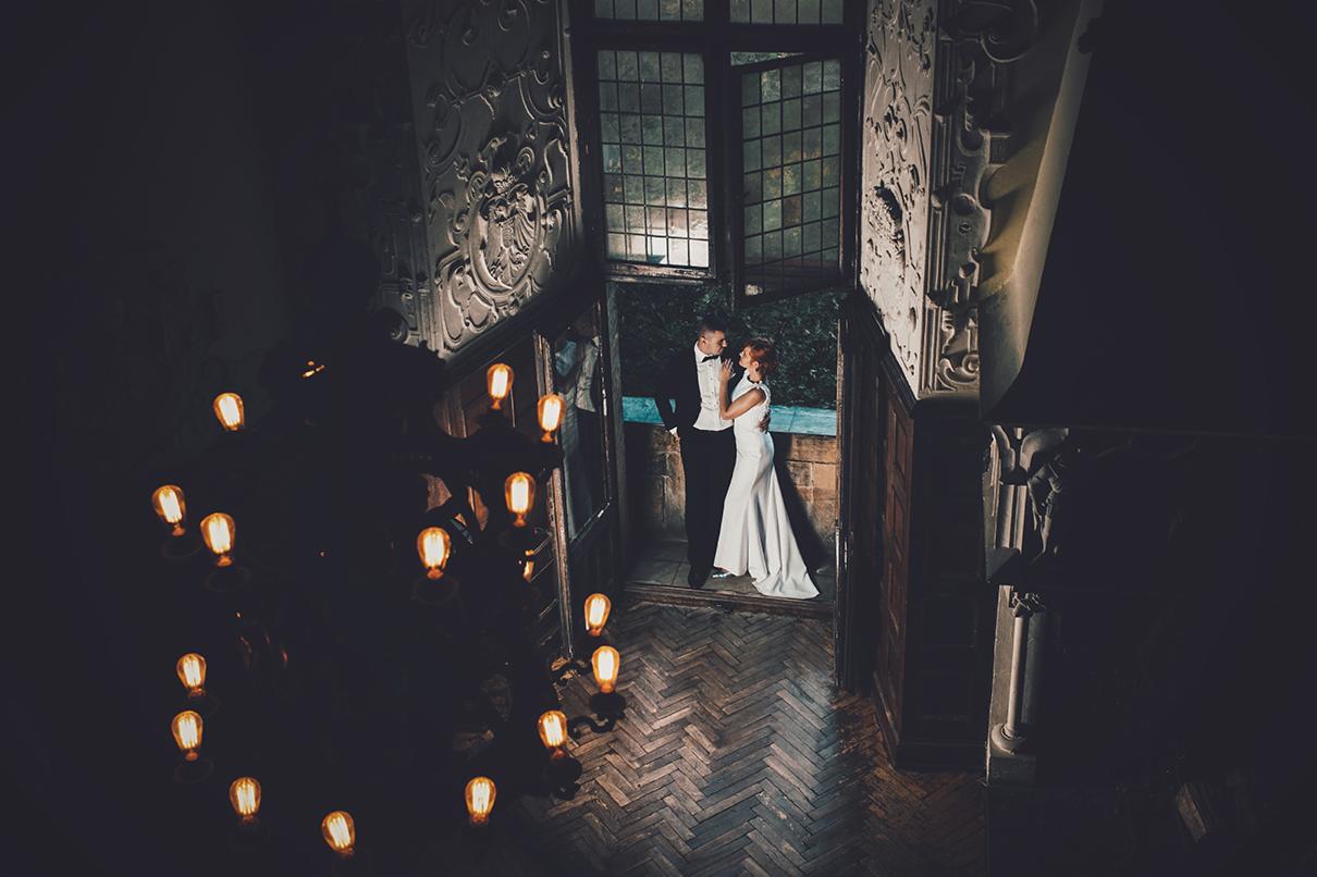 Para młoda w objęciach na tle otwartych drzwi zamku, widok z góry, oświetlenie żarówkami Edisona, dekoracyjny sufit i parkiet w jodełkę.
