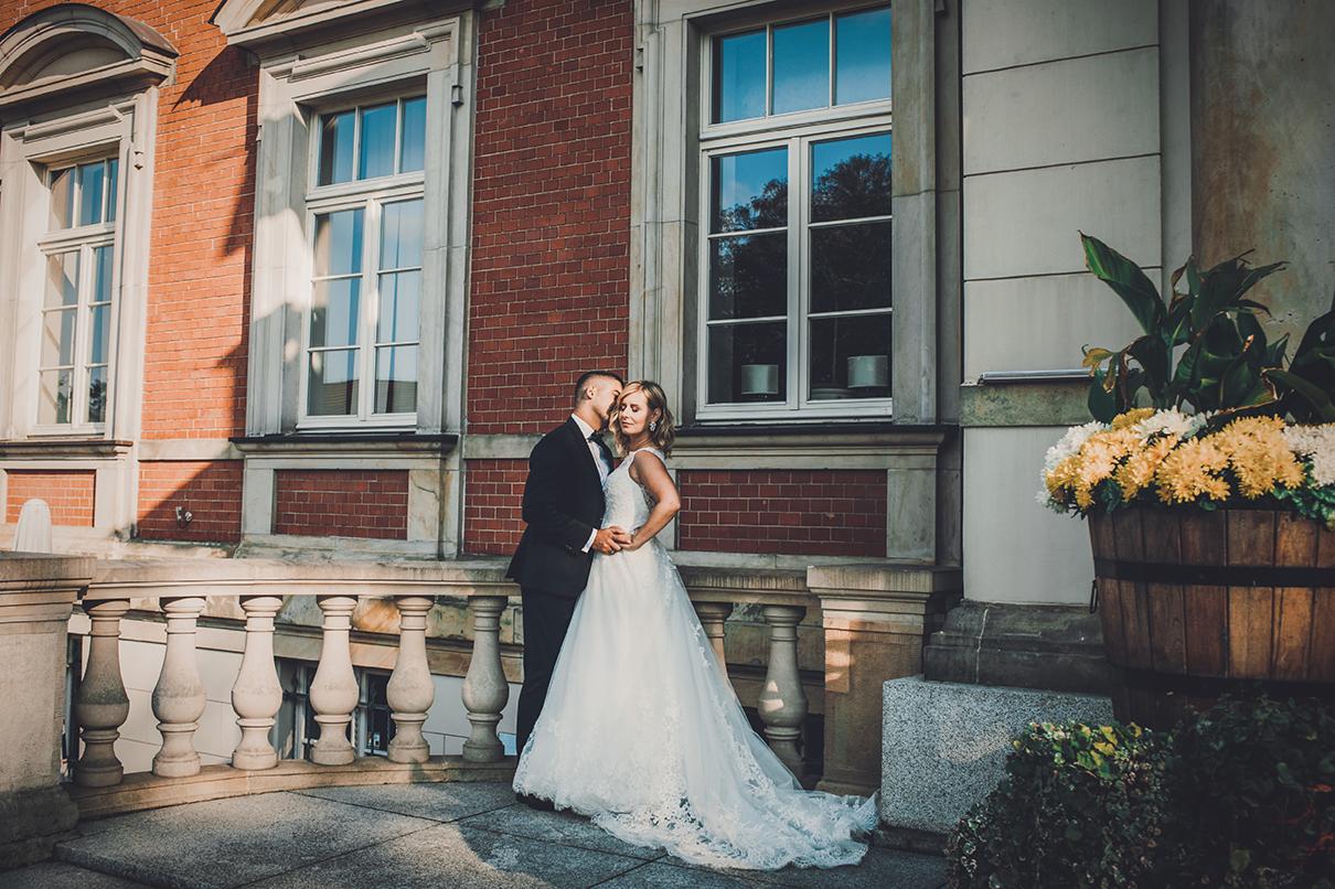 Para młoda w Legnicy pozuje na tle ceglanej fasady budynku z dużymi oknami i kamienną balustradą, obok drewnianej donicy z kwiatami.