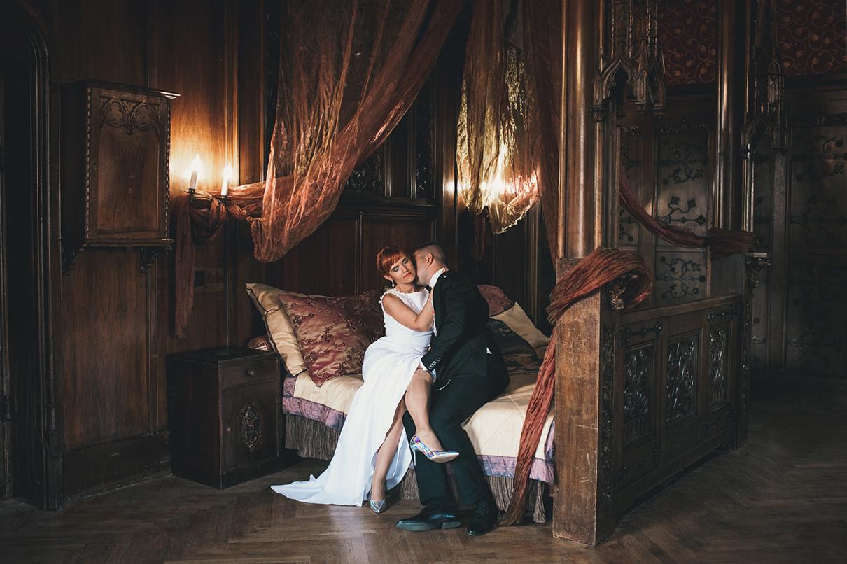 Para młoda siedząca na łóżku z baldachimem w stylu gotyckim, mężczyzna całuje kobietę w policzek, ona ma na sobie białą suknię i srebrne szpilki, on w czarnym garniturze.