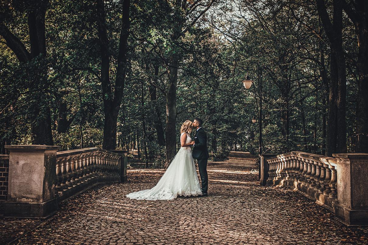 Para młoda całuje się na kamiennym moście w otoczeniu jesiennego parku z latarniami, suknia panny młodej z koronkowym trenem.