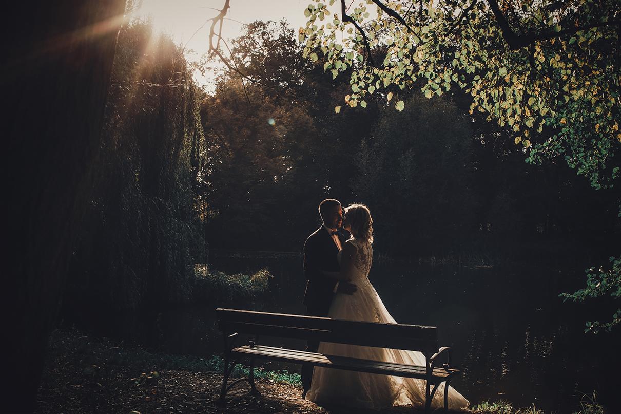 Para młoda w objęciach na tle parku, ławka w pierwszym planie, promienie słońca przebijają przez korony drzew.