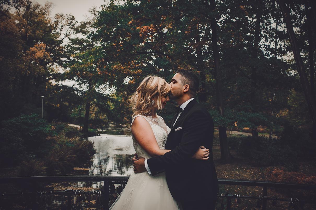 Pan w czarnym smokingu całuje w czoło blondwłosą pannę młodą w koronkowej sukni na tle parku z jeziorem.