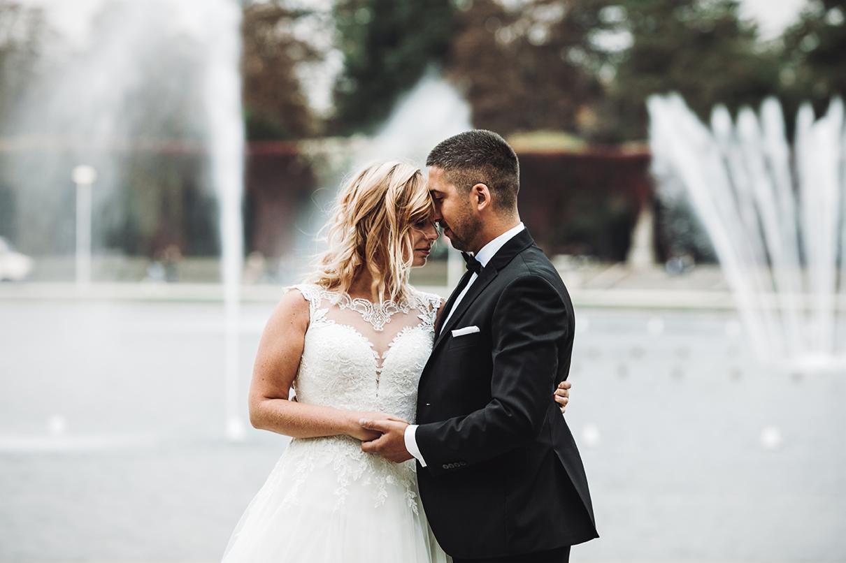 Para młoda, panna młoda w koronkowej sukni i pan młody w smokingu, delikatnie dotykają się czołami na tle fontanny w parku.