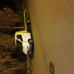 Kris Bud - Biała laweta z żółtym wysięgnikiem zaparkowana nocą na chodniku z kostki brukowej, oświetlona uliczną lampą.