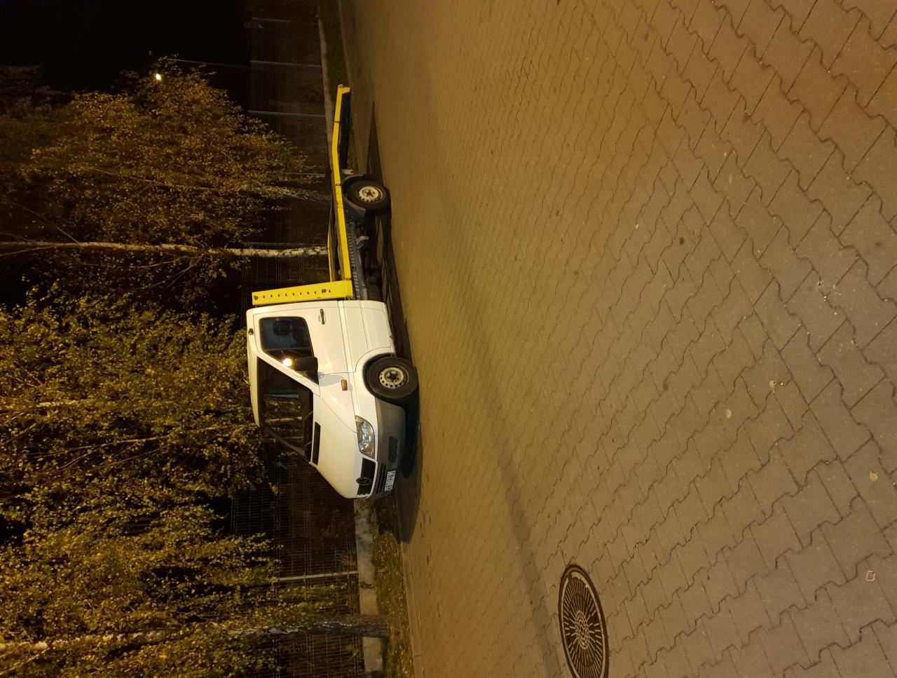 Biała laweta z żółtym wysięgnikiem zaparkowana nocą na chodniku z kostki brukowej, oświetlona uliczną lampą.