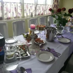 Uroczysty stół z białym obrusem i fioletowymi akcentami, udekorowany kwiatami, świecą, sałatkami i napojami, przygotowany do przyjęcia gości.