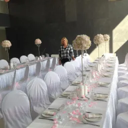 Długi stół weselny z białym obrusem, krzesła w białych pokrowcach zdobione różową wstążką, dekoracje kwiatowe w odcieniach różu, płatki róż na stole, kobieta w tle.