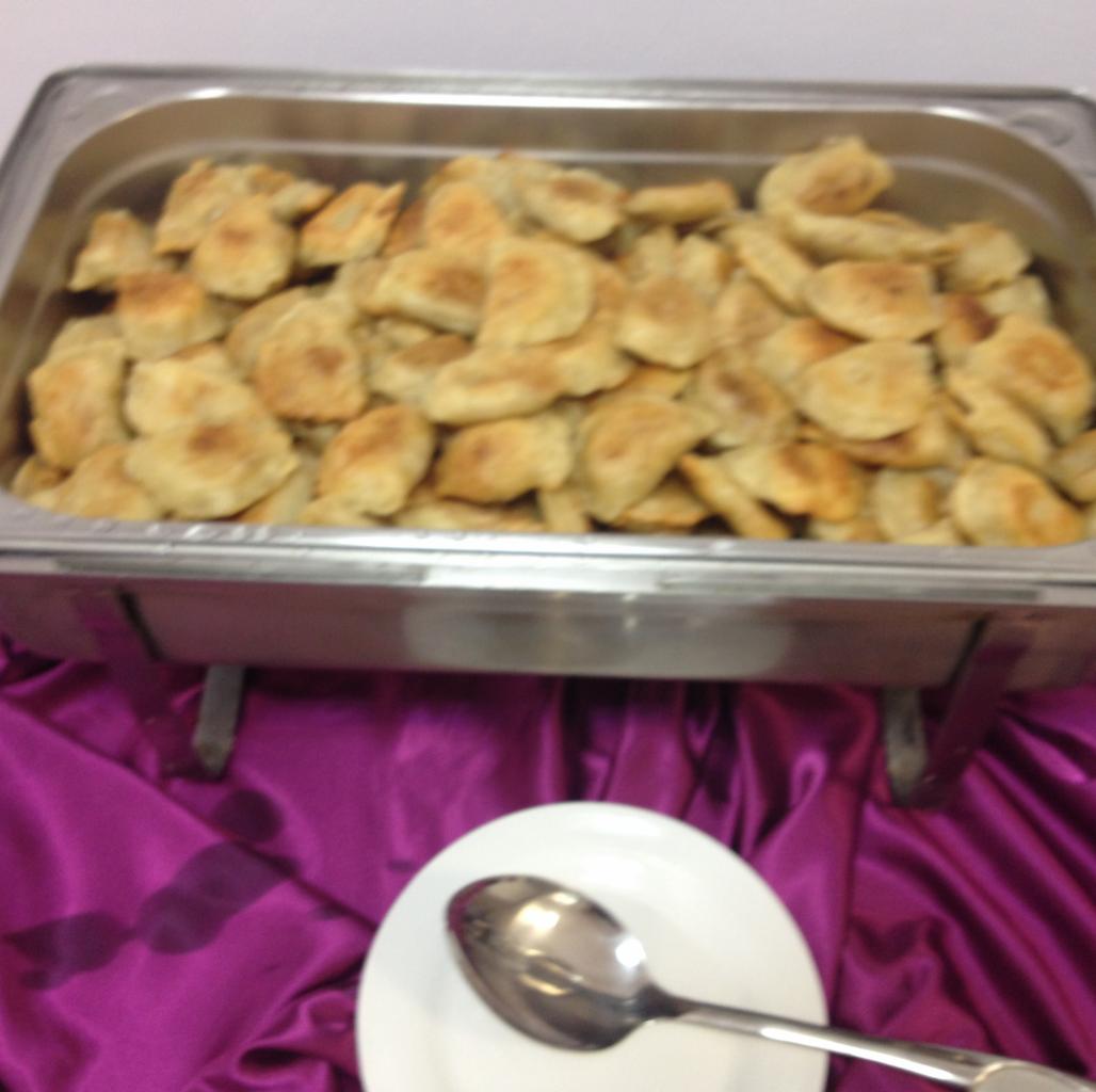 Metalowa podgrzewana taca wypełniona pierogami z farszem, ustawiona na fioletowej, drapowanej tkaninie, z białym talerzem i łyżką w pobliżu.