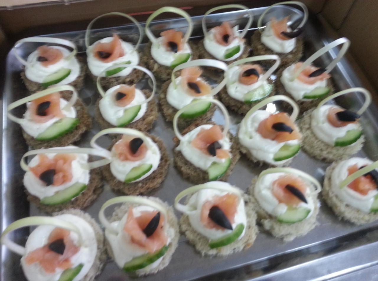 Taca z apetycznymi kanapeczkami: okrągłe pieczywo razowe z kremowym serkiem, plasterkiem ogórka, łososiem i czarną oliwką, udekorowane wstążką z pora.