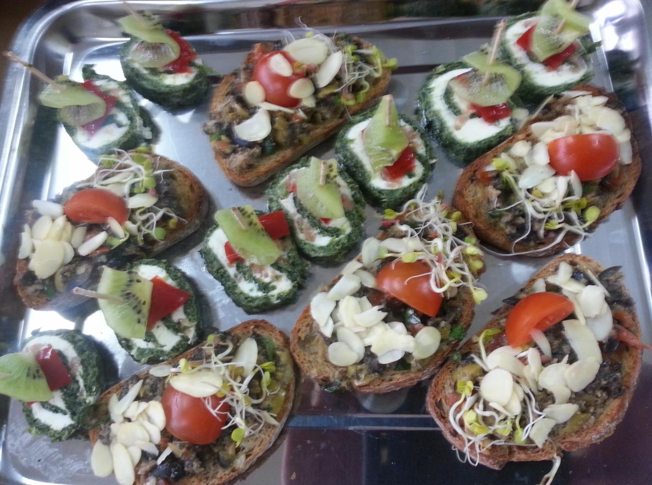 Srebrna taca z różnorodnymi kanapkami: grzanki z pastą warzywną, płatkami migdałów i kiełkami, oraz roladki ze szpinakiem, serem i kiwi, przekrojone na pół i spięte wykałaczką.