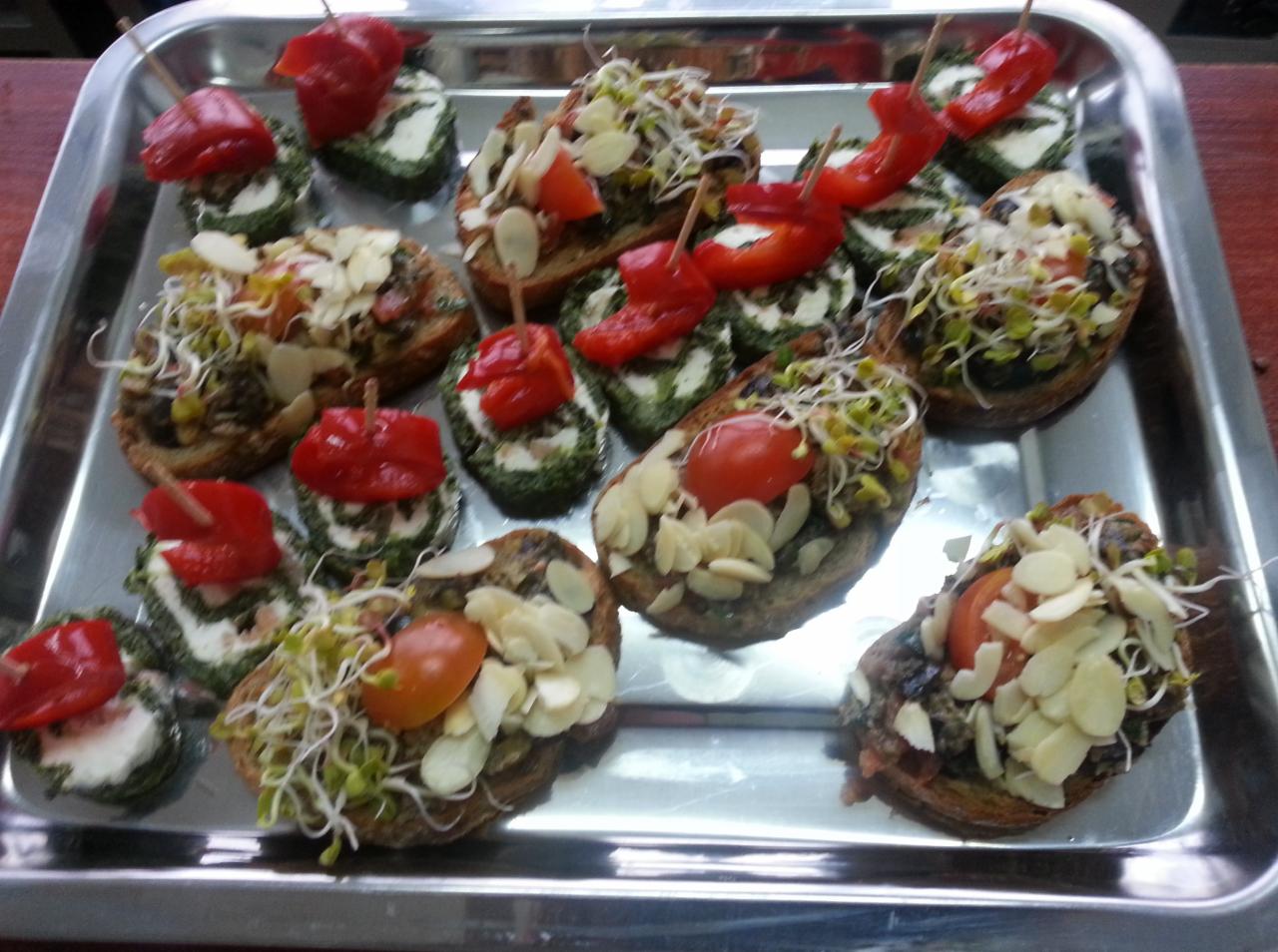 Srebrna taca z różnorodnymi, apetycznymi kanapeczkami: część z kremowym serem i papryką na wykałaczce, inne z pastą, płatkami migdałów i kiełkami, a jeszcze inne z pomidorkiem koktajlowym i kiełkami.
