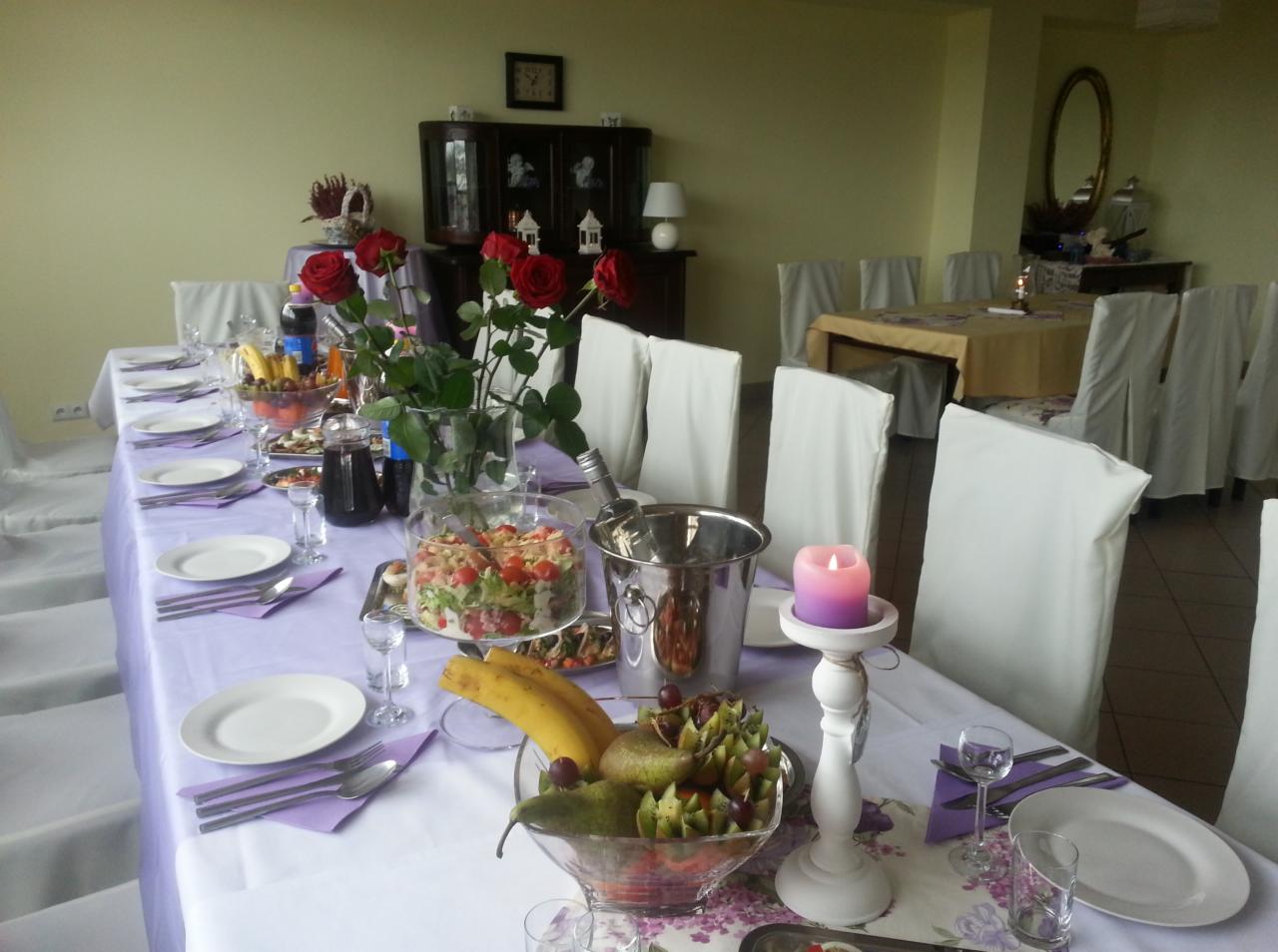 Uroczysty stół z białym obrusem i pokrowcami na krzesłach, dekorowany bukietami czerwonych róż, owocami w szklanych misach, sałatką, napojami w dzbankach, srebrnym wiaderkiem na lód i płonącą...