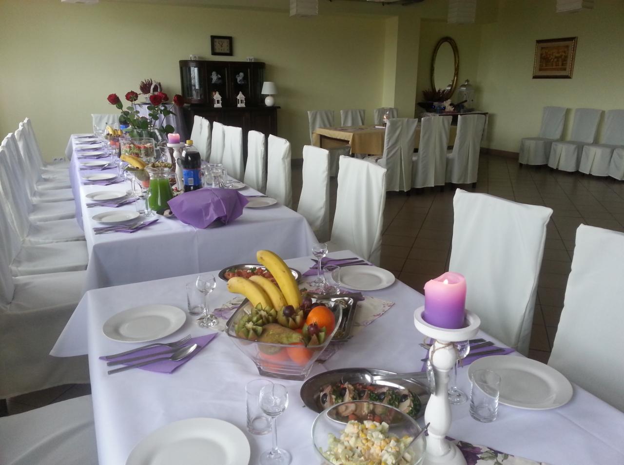 Uroczysta sala bankietowa z długimi stołami nakrytymi białymi obrusami, białymi pokrowcami na krzesłach, dekoracjami z owoców, sałatek, napojów i kwiatów, przygotowana na przyjęcie gości.