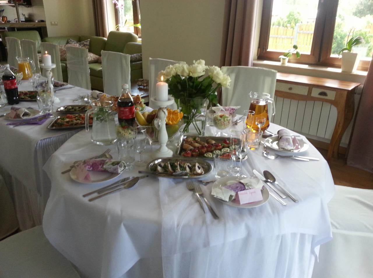 Uroczysty stół z białym obrusem, dekoracjami kwiatowymi, świecami i przekąskami, przygotowany do przyjęcia gości, z widocznym napojem w szklanej butelce z czerwoną etykietą.