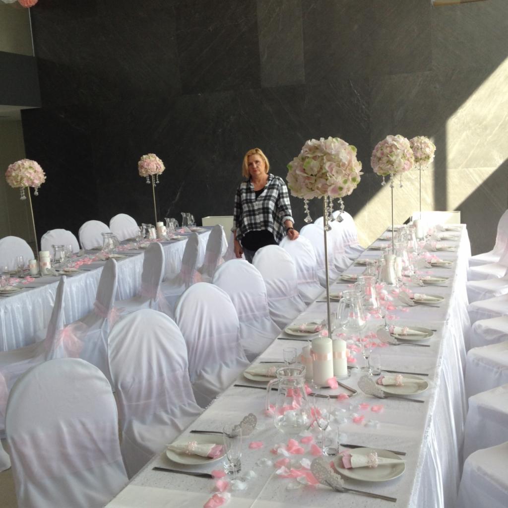 Długi stół weselny z białym obrusem, krzesła w białych pokrowcach zdobione różową wstążką, dekoracje kwiatowe w odcieniach różu, płatki róż na stole, kobieta w tle.
