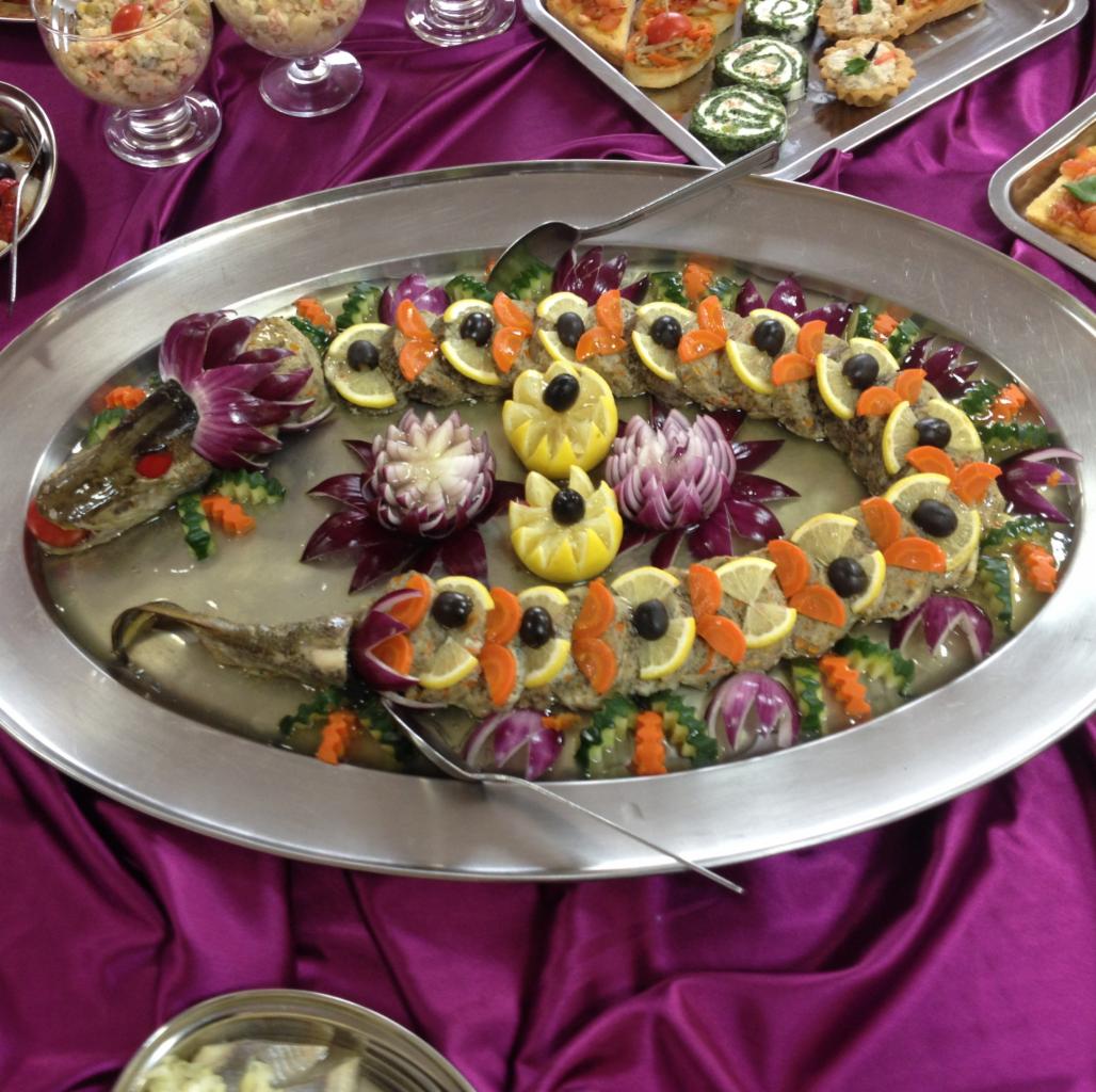 Dekoracyjna potrawa rybna ułożona w kształt węża na srebrnej tacy, ozdobiona cytrynami, oliwkami, marchewką i fioletową cebulą, podana na fioletowym obrusie.