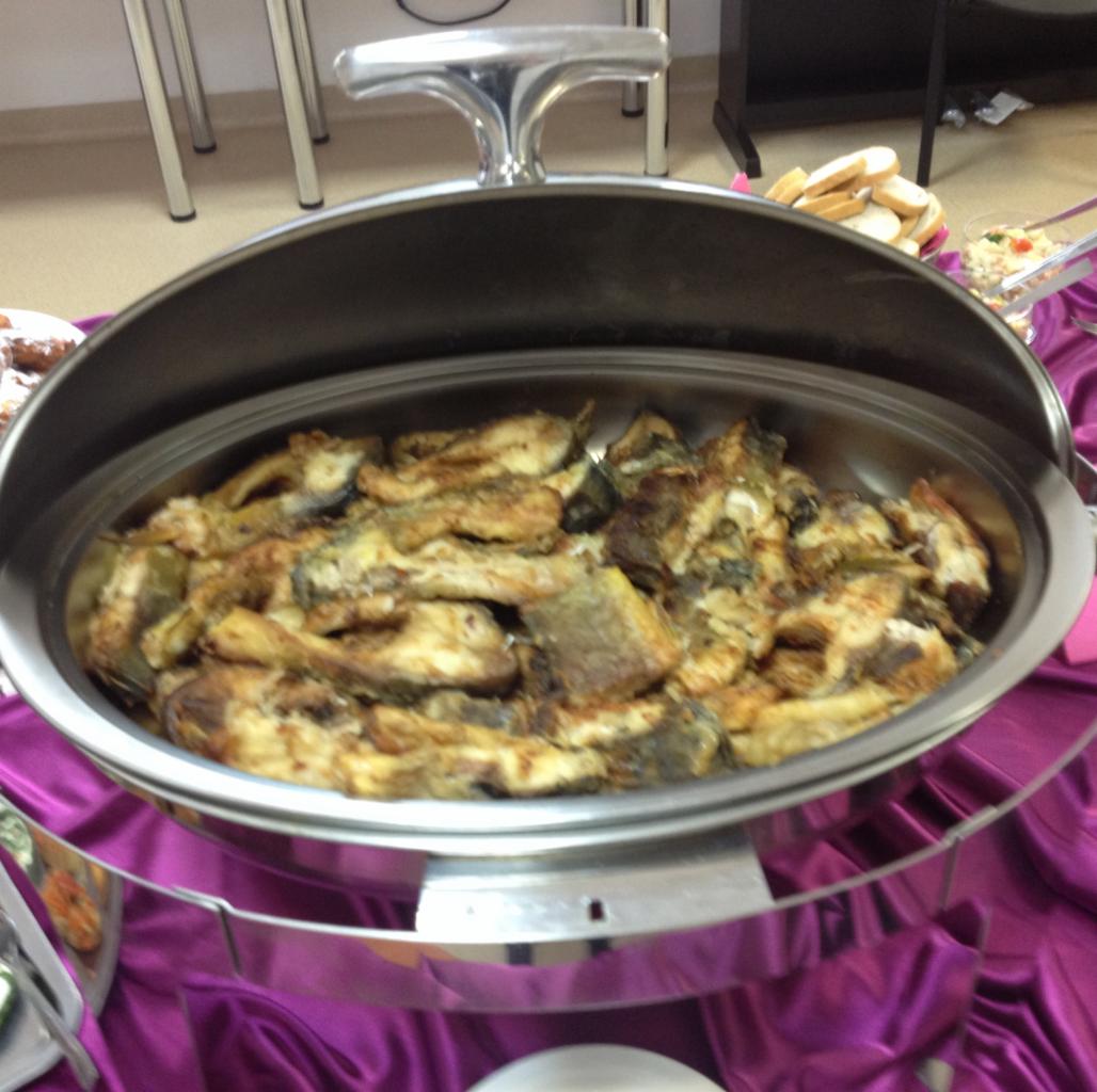 Podgrzewacz bufetowy ze smażonymi kawałkami ryby na fioletowym obrusie, widoczne pieczywo i sałatka w tle.