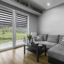 Nowoczesny salon z dużymi oknami przesuwnymi, wyposażonymi w rolety dzień-noc, widok na zielony ogród, minimalistyczny wystrój wnętrza.