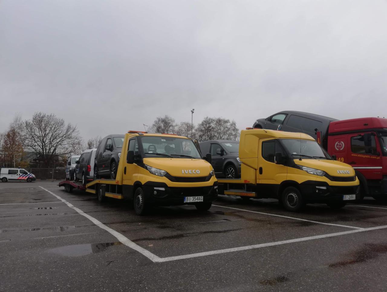 Dwie żółte lawety Iveco na parkingu; jedna z trzema samochodami osobowymi na platformie, druga z jednym busem; w tle czerwona ciężarówka i radiowóz policyjny.