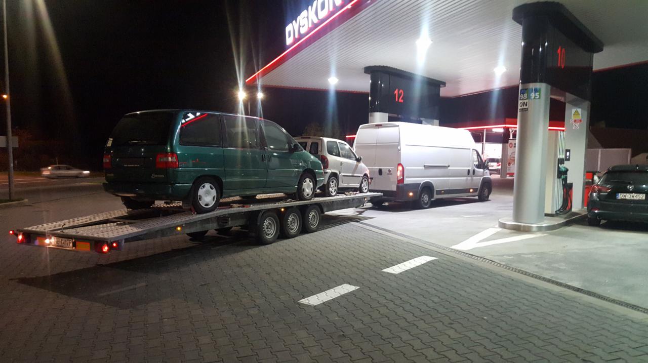 Dwa samochody osobowe, w tym zielony van z tyłu, umieszczone na platformie lawety, ciągniętej przez białego busa na stacji benzynowej oświetlonej neonami w nocy.