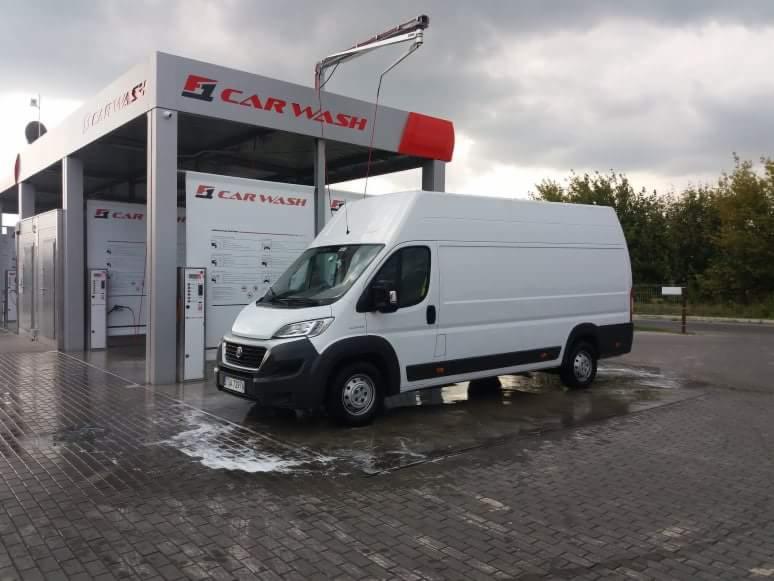 Biały bus dostawczy po umyciu w samoobsługowej myjni samochodowej, widoczne ślady piany na kostce brukowej.