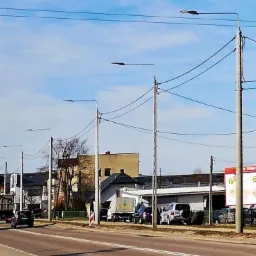 Ulica miejska z rzędem słupów oświetleniowych, widoczne samochody i budynki w tle, pochmurne niebo.