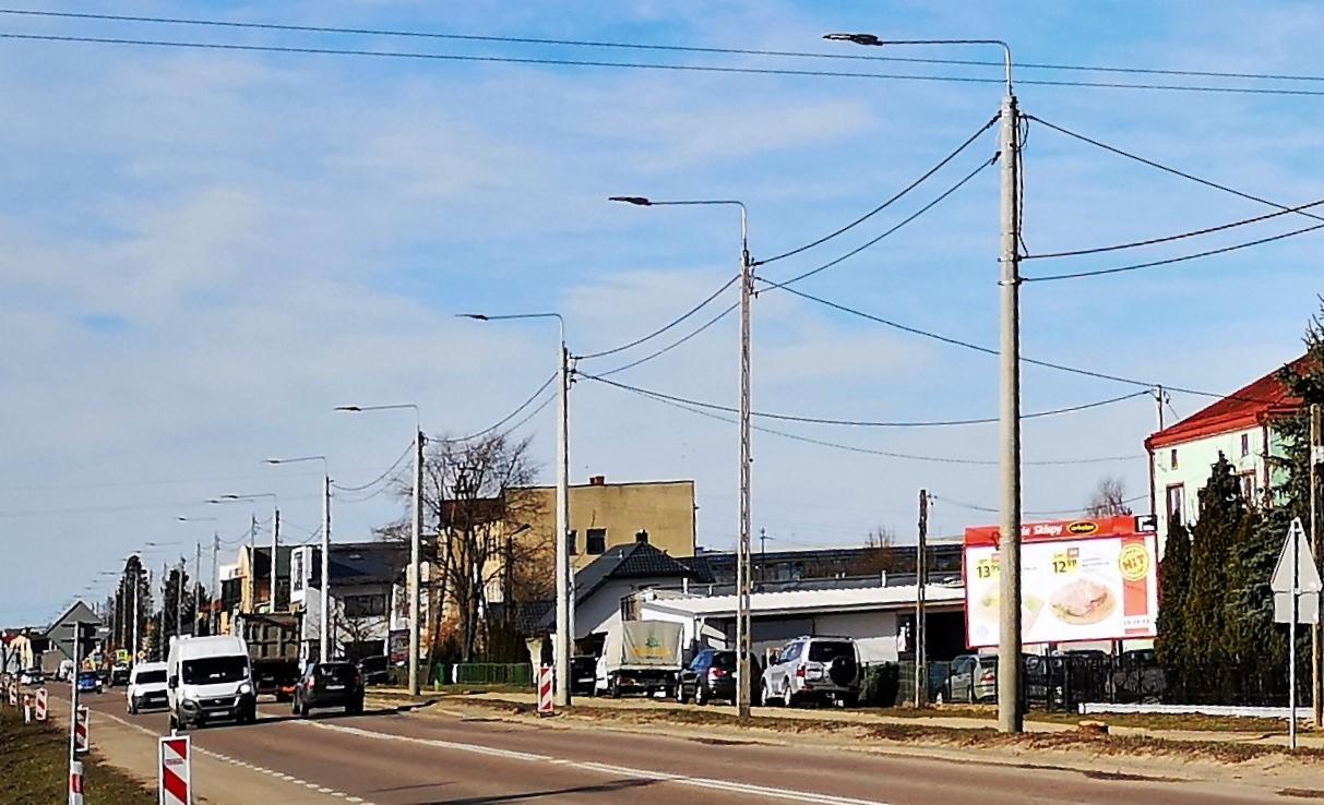 Ulica miejska z rzędem słupów oświetleniowych, widoczne samochody i budynki w tle, pochmurne niebo.