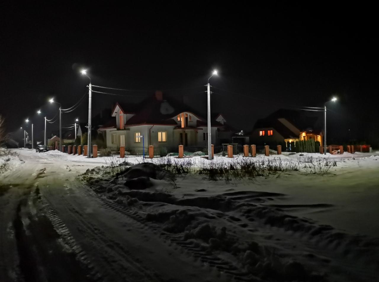 Zimowy krajobraz nocą z oświetlonymi domami i ulicznymi lampami LED, widoczne ślady opon na śniegu.