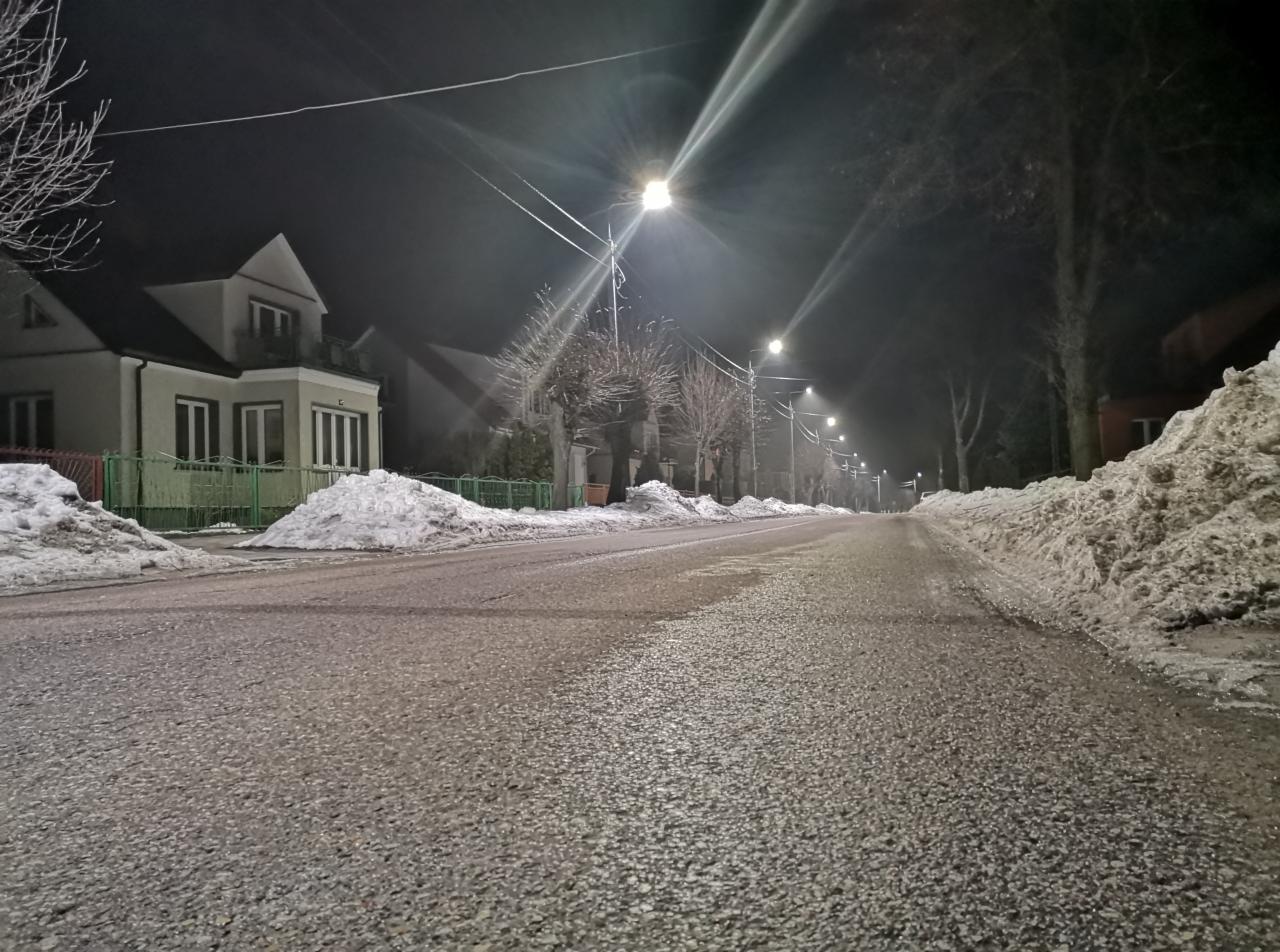 Ulica oświetlona przez latarnie uliczne nocą, z odśnieżonymi poboczami i widocznymi domami w tle.