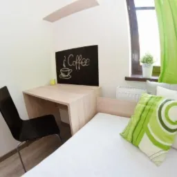 Pokój z biurkiem, krzesłem, łóżkiem jednoosobowym, zielonymi zasłonami i tablicą z napisem 'Coffee' narysowanym kredą.