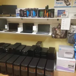 Sklep z elektroniką: Rząd czarnych komputerów stacjonarnych na podłodze, laptopy i monitory na półkach, akcesoria komputerowe i drukarki w tle, certyfikat Farex widoczny na górnej półce.