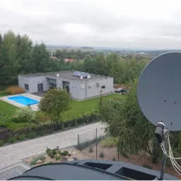 Widok z dachu na nowoczesny dom z basenem i panelami słonecznymi, przesłonięty anteną satelitarną na pierwszym planie.